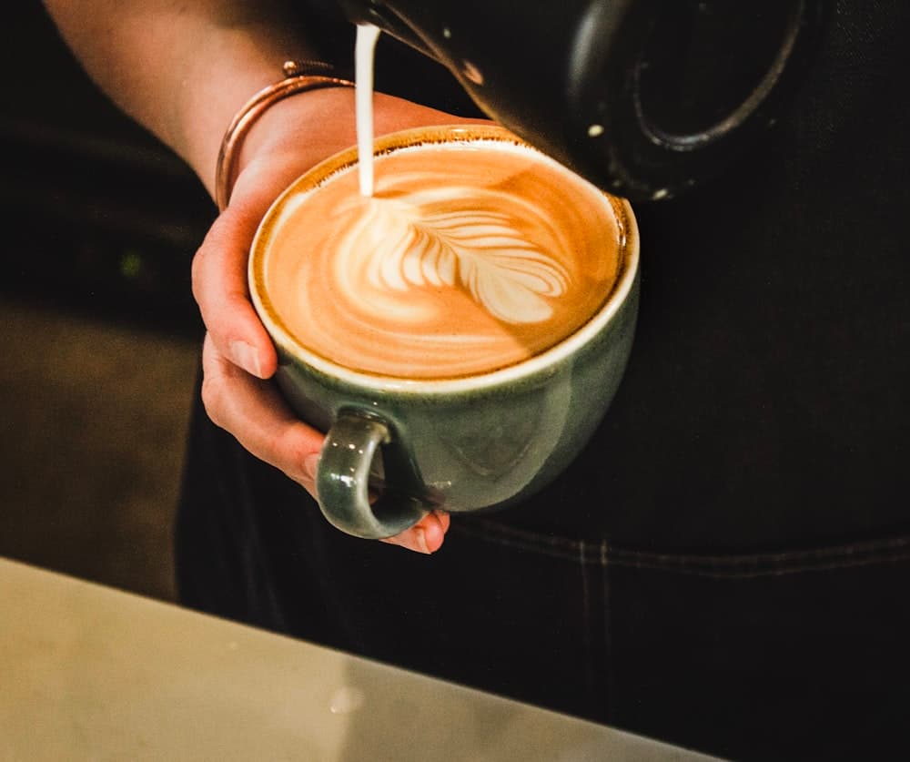 Tangan menuang susu ke cangkir keramik hijau membentuk latte art
